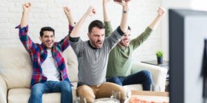 Three men sit on a couch with their arms raised in a cheering position.