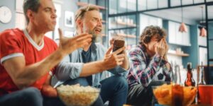 Three men sit across a couch with popcorn on a table in front of them. They all look frustrated with the game they are watching.