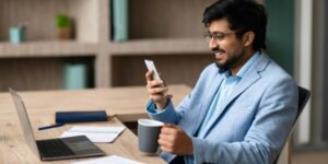 A man sits at a desk with a cup of coffee, watching a video on a smartphone.