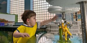A young boy in a yellow smock reaches for water pouring out of a play feature.