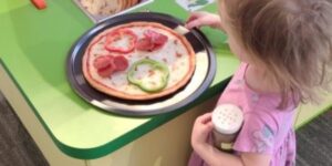 A young girl makes a pretend pizza at a green counter.
