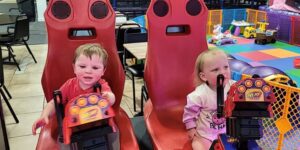 A toddler boy and girl sit in red seats at a play center.