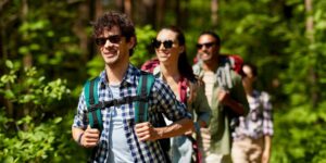 A group of young adults wearing rugged clothing and backpacks walk outside near some woods.
