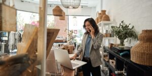 A woman stands at a counter or desk in a boutique shop, talking on the phone and looking at a laptop.
