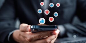 Close-up of hands holding a smartphone. "Like" and "heart" icons float above the phone.