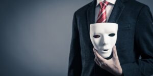 A man in a business suit with a red tie holds a blank drama mask in one hand.