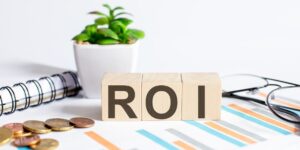 The letters "ROI" in black ink on wood blocks sit on a desk by a notebook and a potted plant.