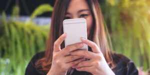 A woman in the background holds her smartphone in the foreground with both hands.