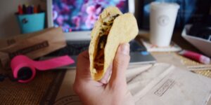 A hand holds a taco in front of a computer monitor.