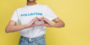 A woman in jeans and a white shirt with the word "volunteer" written in blue lettering makes a heart out of her hands.