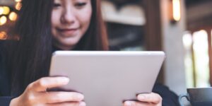 A young woman with long dark hair holds a tablet in both hands.