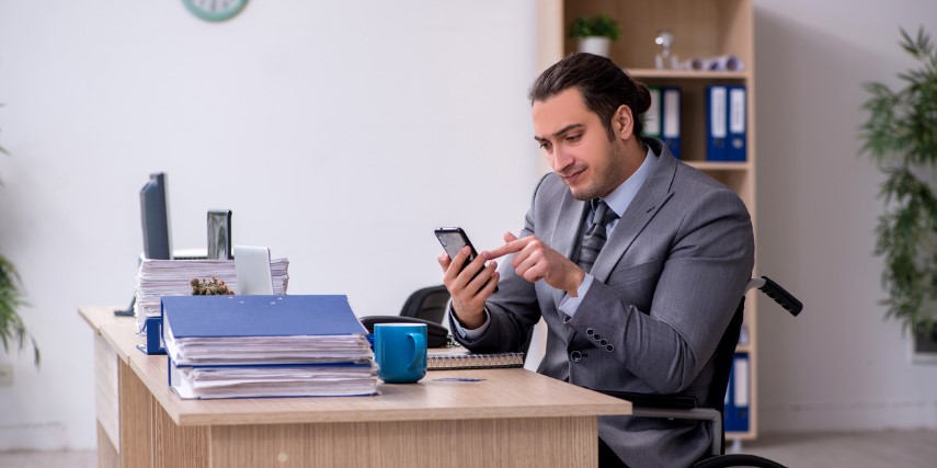 A man in a wheelchair sits at a desk scrolling on a smartphone.