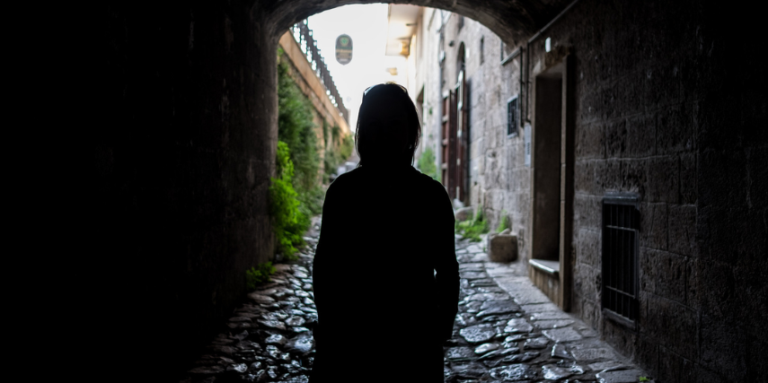 A person in a hoodie stands in a dark tunnel looking out at the light at the end of the tunnel.