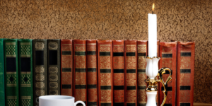A row of old-fashioned hardback, cloth-bound books with a teacup and a lit candlestick in front of them.