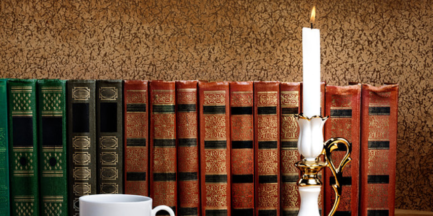 A row of old-fashioned hardback, cloth-bound books with a teacup and a lit candlestick in front of them.