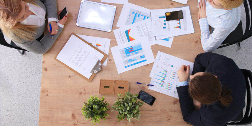 Overhead view of three people sitting at a table with printed charts and graphs scattered on the surface.