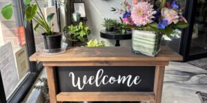 A wooden stand with a black sign that says, "Welcome," and has a flower arrangement on top.