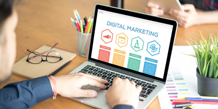 A person sits typing on a laptop with the words "Digital Marketing" and several graphics on the screen in vibrant colors.
