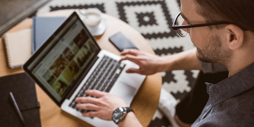 Close-up of a laptop on a wood table and a bearded man with glasses working on creating content.