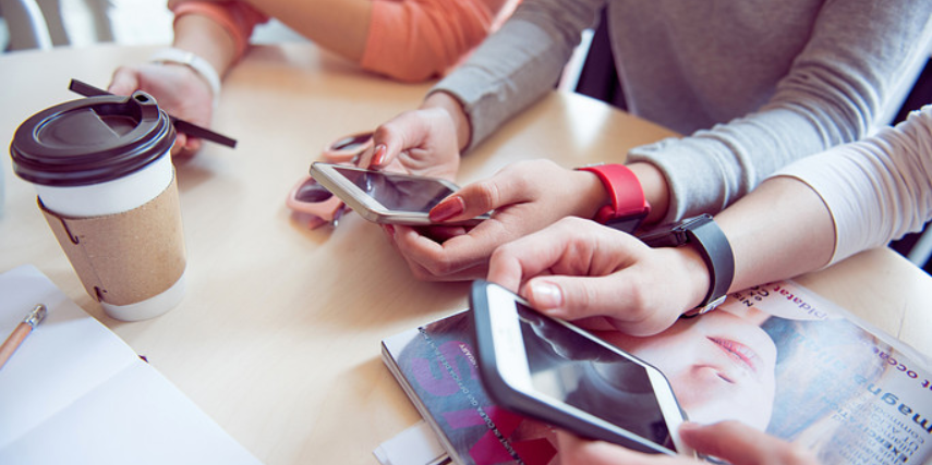 A group of friends gather at a table, all looking at their phones while enjoying coffee.