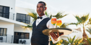 A waiter is outside, carrying two orange and yellow cocktails on a tray.