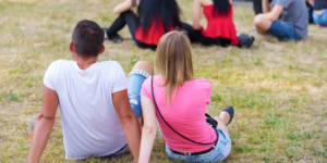 A man and woman are seen from behind, sitting in the grass at an outdoor music event.