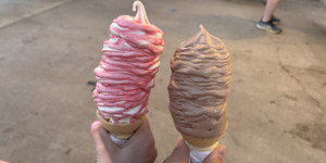 Close-up of two hands holding two large soft serve ice cream cones.