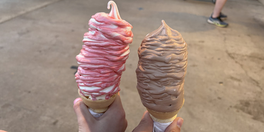 Close-up of two hands holding two large soft serve ice cream cones. 