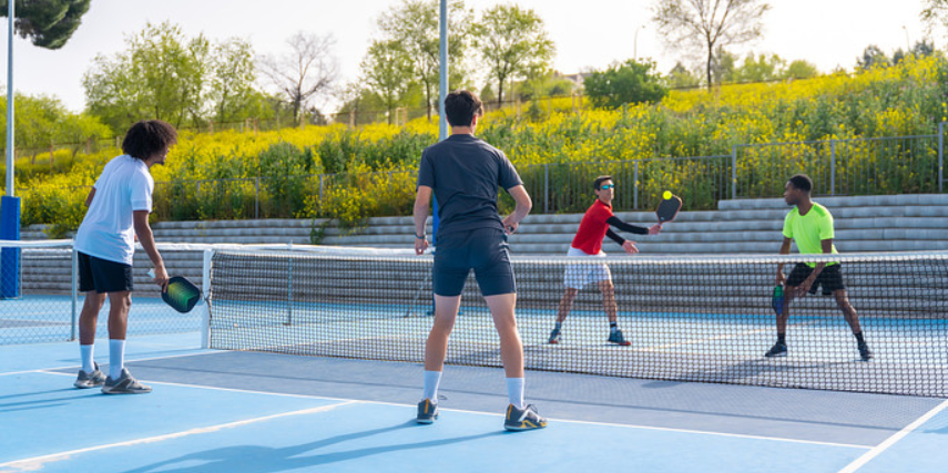 Four people play pickleball on a court surrounded by greenery.