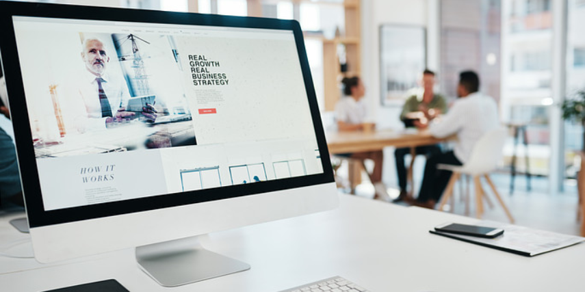 A desktop monitor on a white desk in a busy office shows a business website.