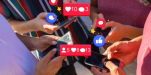 A group of people stands together looking at their phones. Social media icons float above their phones.