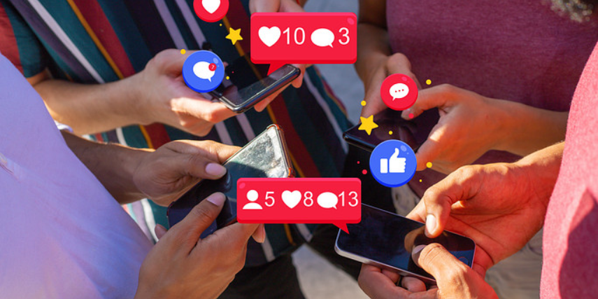 A group of people stands together looking at their phones. Social media icons float above their phones.