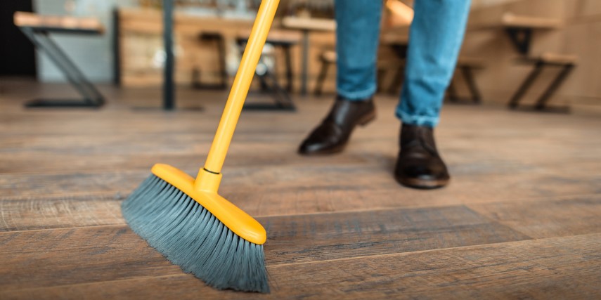 A yellow and gray broom sweeps across a wood floor with a person behind it presumably holding the broom. 