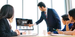 People are gathered around a table at a meeting while one man stands, pointing at a monitor with the word "Blog" across it.