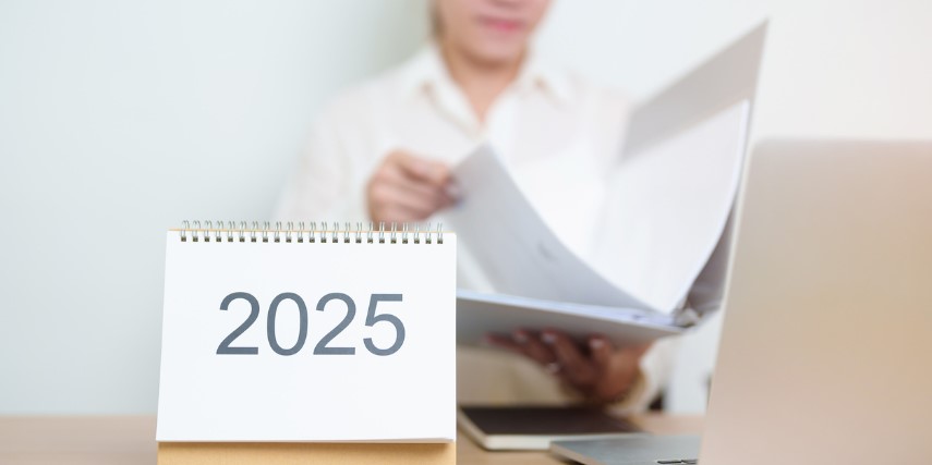 A woman sits reviewing a report at her desk with a laptop and a flip chart that says, "2025" in black against a white background.