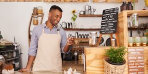 A young man wearing an apron stands at the counter of a coffee shop looking at his phone.