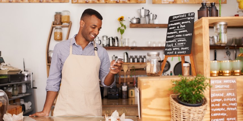 A young man wearing an apron stands at the counter of a coffee shop looking at his phone.