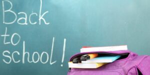 An open purple backpack against a chalkboard where "Back to School!" is written in white chalk.
