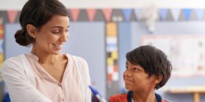 A female teacher and a young male student sit in a classroom, smiling at one another.