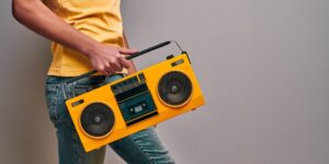 Man in a yellow shirt and blue jeans carries a bright yellow boom box in front of a white wall.