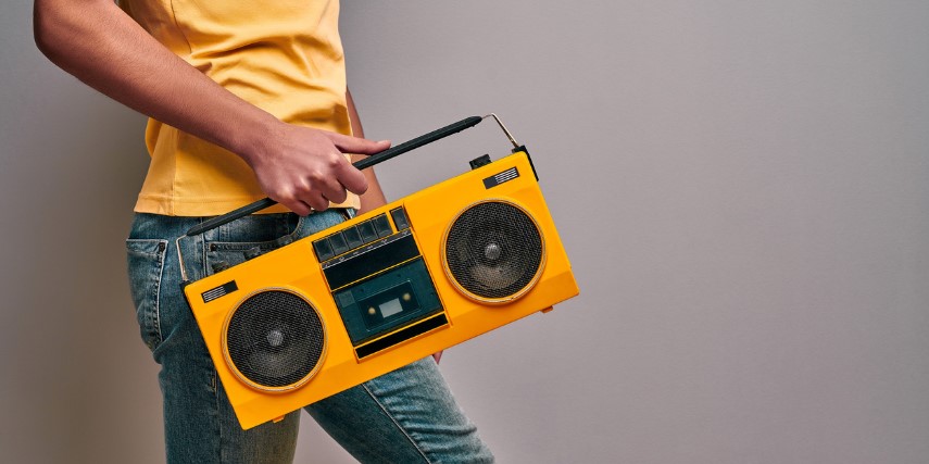 Man in a yellow shirt and blue jeans carries a bright yellow boom box in front of a white wall.