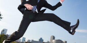 A man in a black business suit and shiny black shoes jumps in front of a city skyline.
