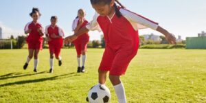 A young girl in a red uniform kicks a soccer ball while other players run toward her in the background.