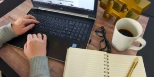 A laptop, spiral notebook and a cup of coffee sit on a wood table. A woman is poised to type on the laptop.