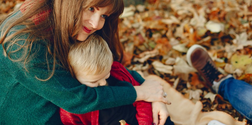 A mom with long, dark hair snuggles her son in a pile of leaves.