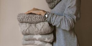 A woman in a gray sweater with a silver bracelet holds a stack of folded gray sweaters.