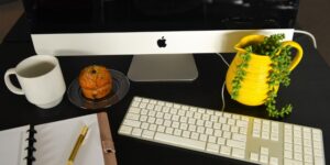 Overhead view of a desk with a desktop computer, muffin, plant, cup of coffee and notebook.