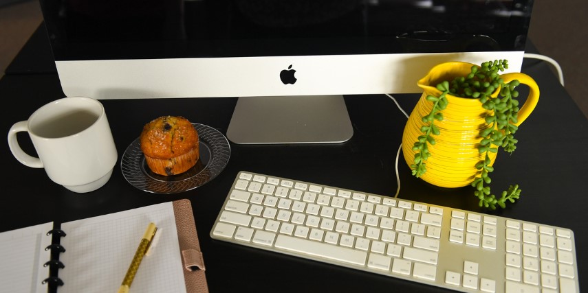 Overhead view of a desk with a desktop computer, muffin, plant, cup of coffee and notebook.