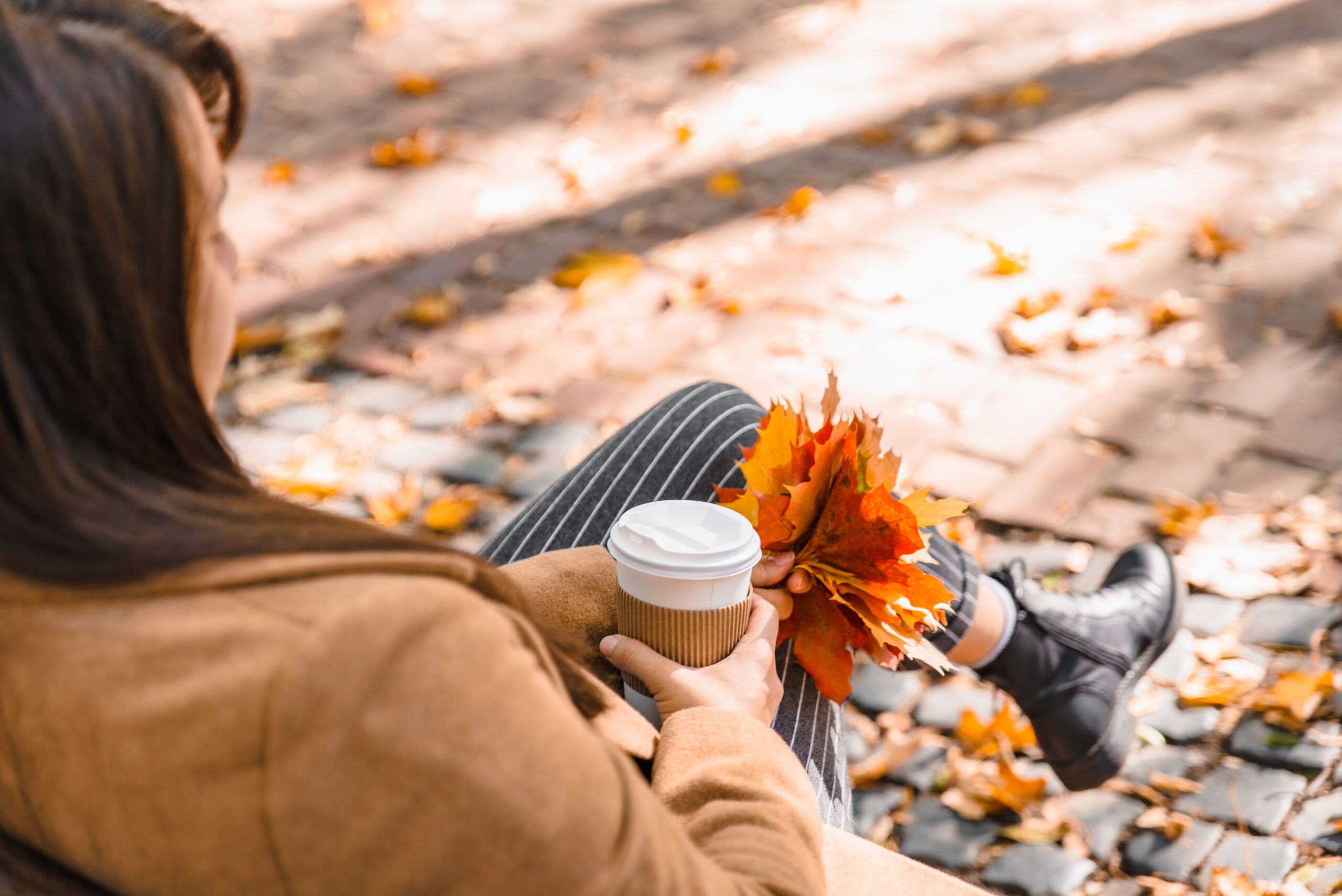 Girl holding coffee cup and leaves while sitting on bench in the fall. 