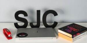 A laptop, red stapler, two books and a large "SJC" in black letters are displayed on a white surface.